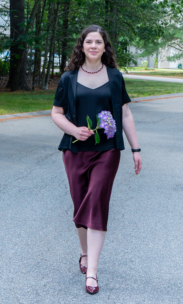 Woman walking toward camera on a neighborhood street, holding a purple hydrangea bouquet and wearing a black top, blazer, burgundy skirt, red shoes, and a beaded necklace.