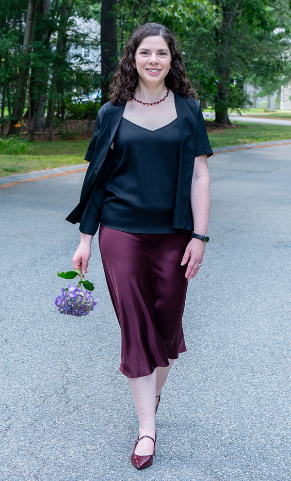 Smiling woman walks along a tree-lined street in a black blazer and burgundy outfit, holding a small bouquet of purple flowers.
