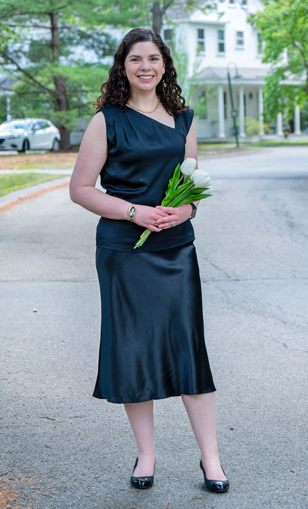 Woman in a navy satin dress standing on a residential street, smiling, and holding a bouquet of white tulips.