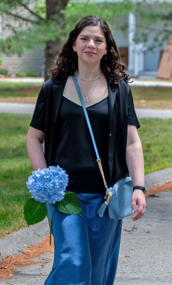 Woman walking along a campus path holding a light blue hydrangea, dressed in a black top, blue satin pants, and a matching light blue purse.