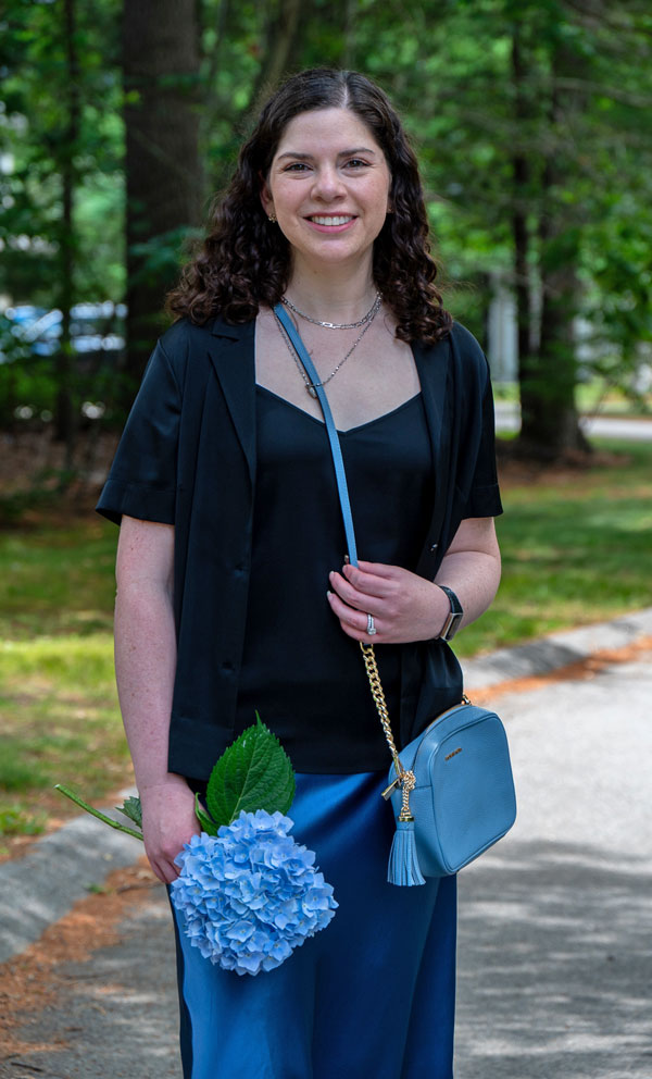 Smiling woman outdoors in a park, wearing a dark blazer and blue outfit, holding a blue hydrangea bouquet and a blue purse with tassel.