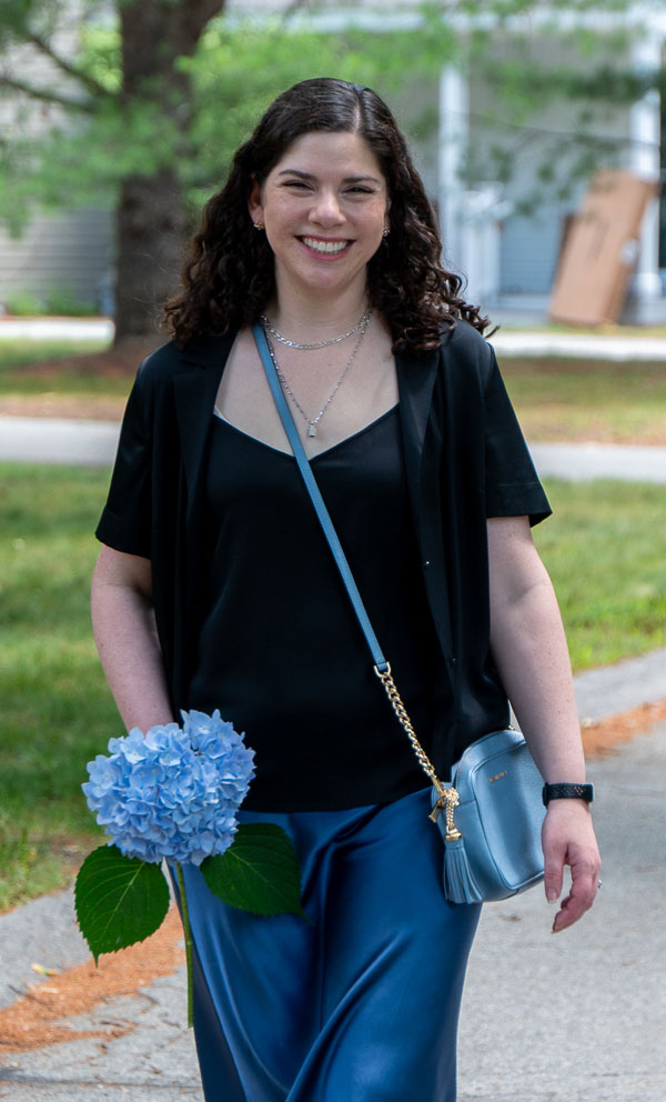 Smiling woman walking outdoors holding a blue hydrangea, wearing a black top, blue pants, and a light blue crossbody bag.