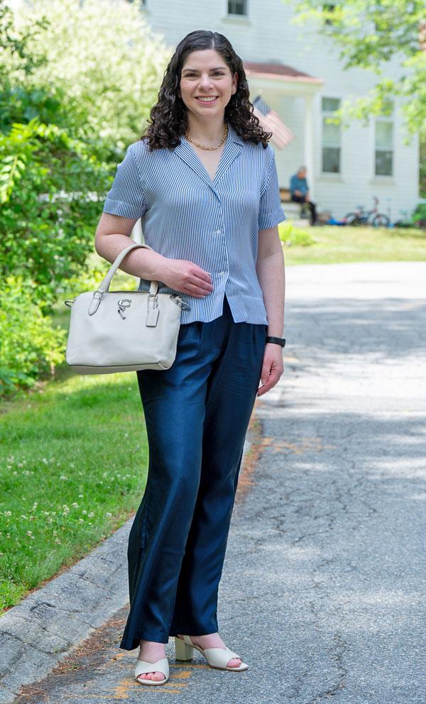 Smiling woman standing on a suburban street with a beige handbag, wearing a blue striped blouse and navy pants.