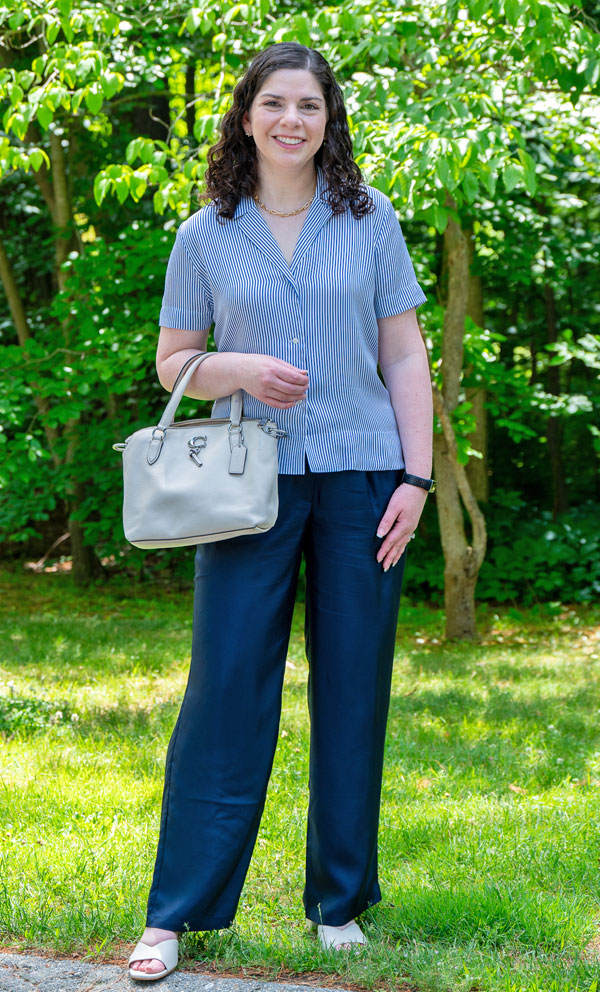 Woman smiling while standing outdoors in a garden, wearing a blue striped short-sleeve blouse, navy wide-leg pants, white sandals, and carrying a light gray handbag.