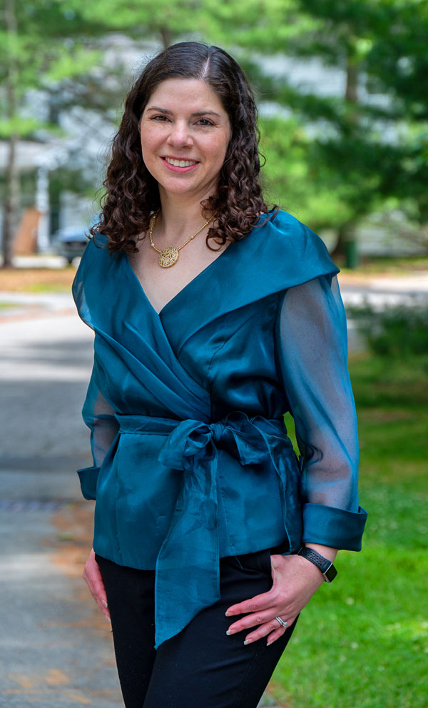 Smiling woman with curly brown hair wearing a teal wrap blouse and gold pendant, standing on a tree-lined path outdoors.