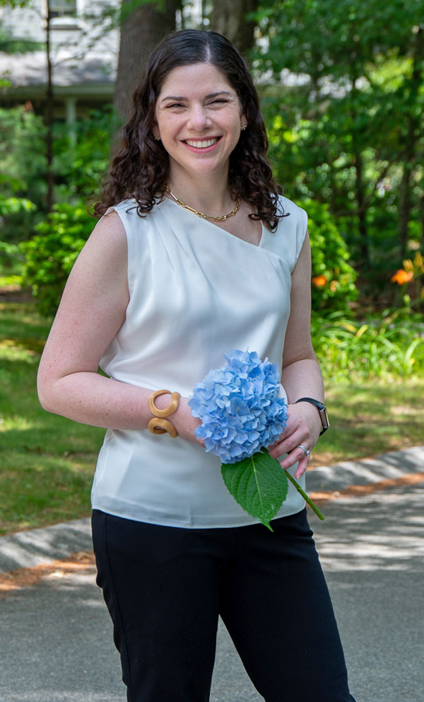 Smiling woman outdoors in a white sleeveless blouse, holding a blue hydrangea with green leaves, in a sunny garden setting.