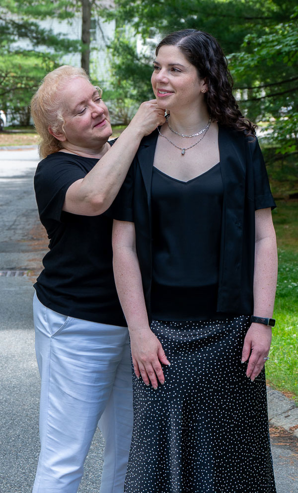 Older woman adjusts a necklace on a younger woman outdoors, both wearing black tops.