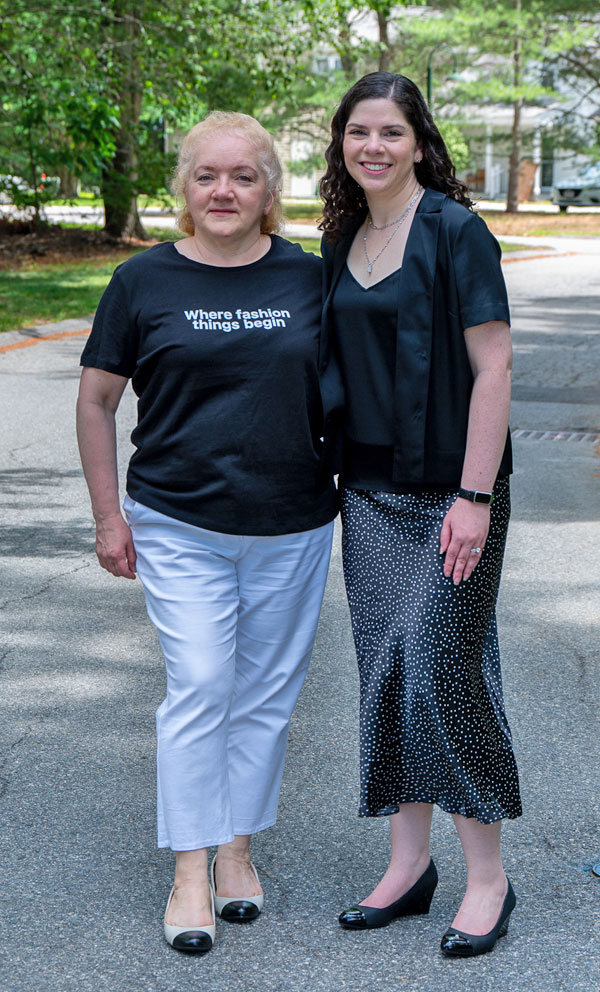 Two women posing together outdoors on a sunny street; left woman in a black T‑shirt that reads 'Where fashion things begin' and white pants, right woman in a dark blazer and dotted skirt, both smiling.