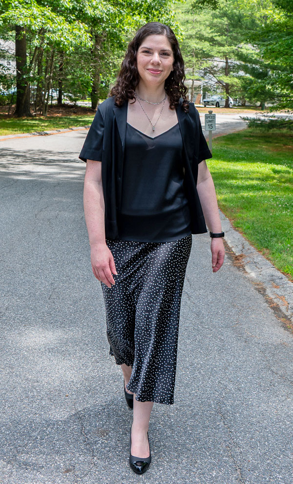 Smiling woman walks toward the camera on a sunlit, tree-lined street, wearing a black blazer, silky black top, and dotted wide-leg pants.