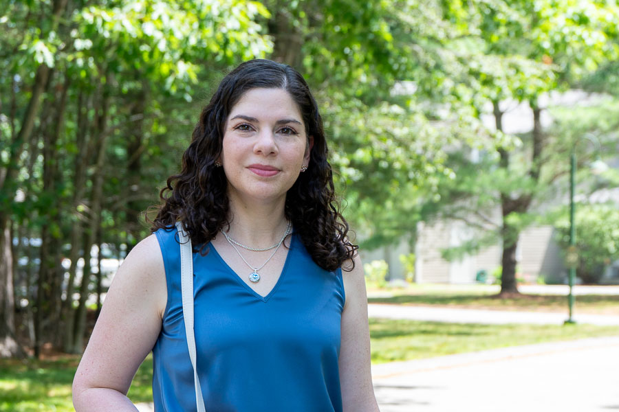 Woman with curly dark hair in a blue sleeveless top standing on a sunny park path with trees behind her.